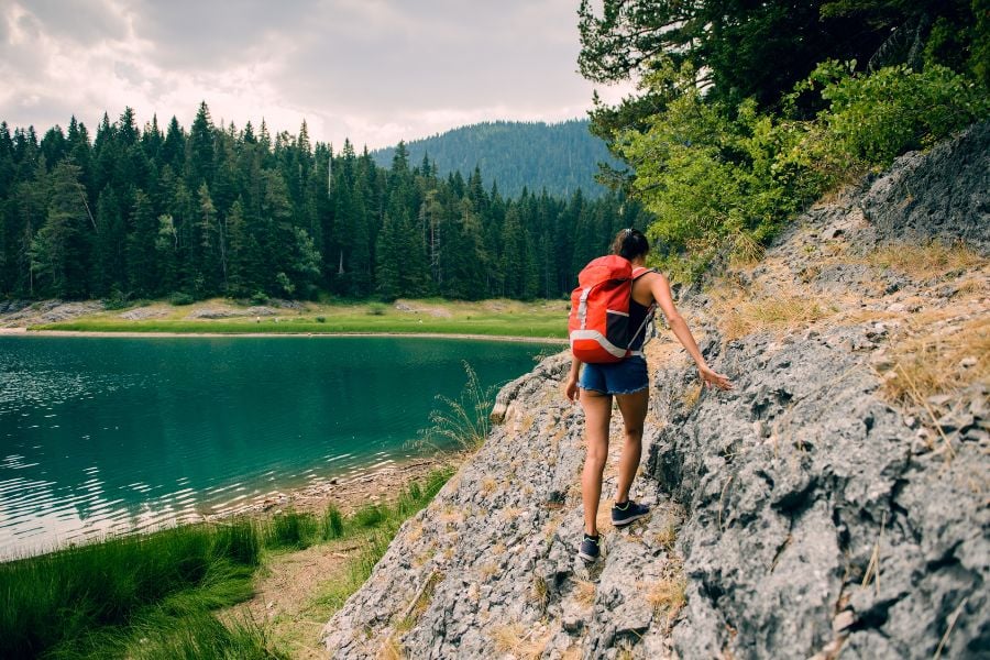 A hiker with a red backpack climbs rocky terrain beside a turquoise lake.