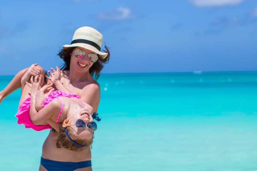 A smiling mother twirls her young daughter on a bright, tropical beach with crystal-clear water.