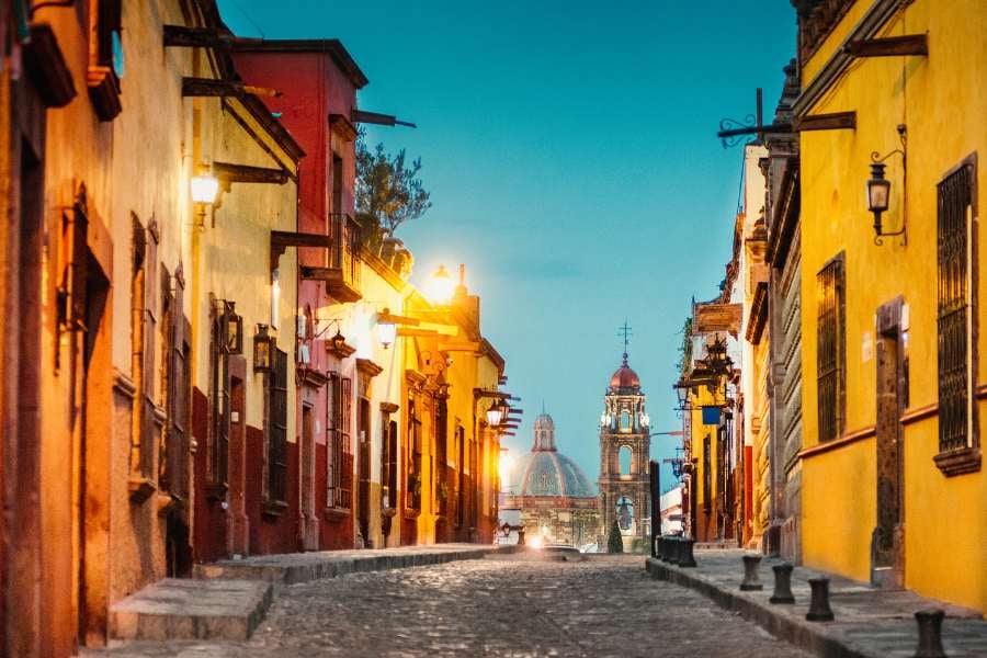 A charming cobblestone street glows under warm streetlights, leading to a historic church dome in the distance.