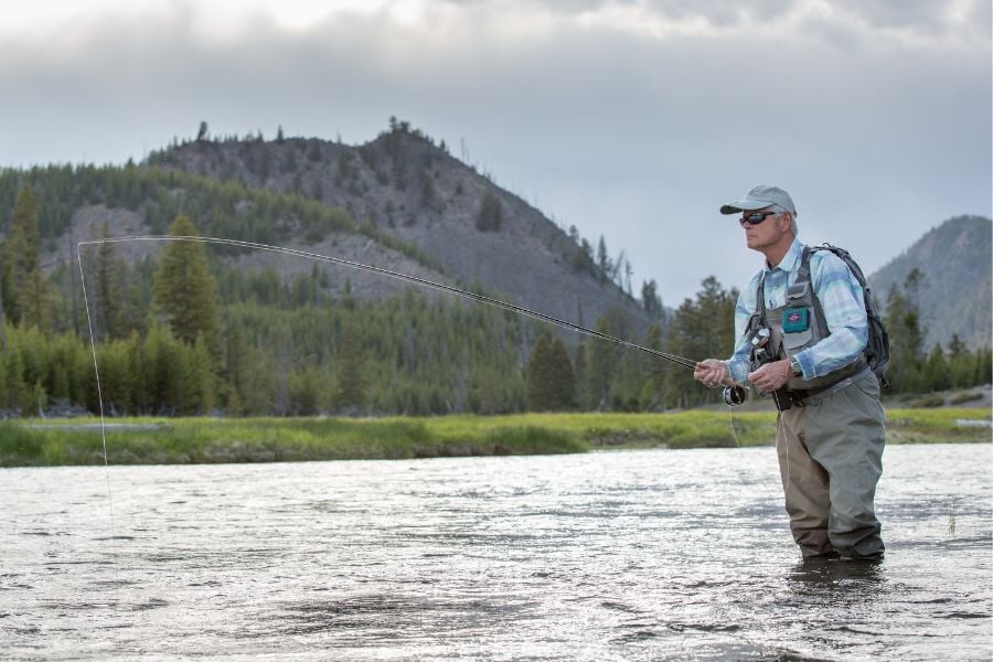 Fisher stands knee-deep in Yellowstone river, casting steadily in quiet waters.