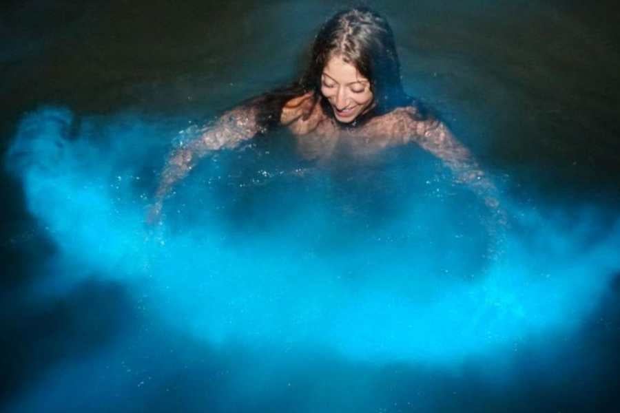 A woman floats in glowing blue water, surrounded by magical bioluminescent light in a dark and peaceful lagoon.