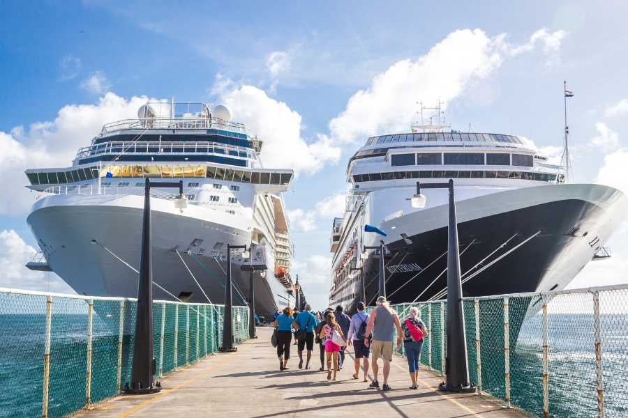 A bustling cruise pier with passengers walking between two massive ships on a sunny day.