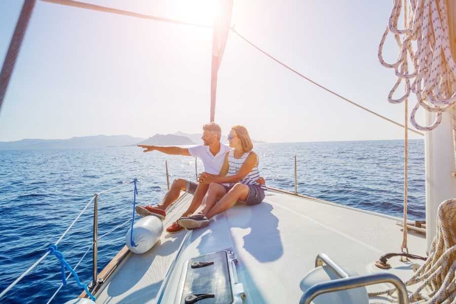 A couple sits at the bow of a sailboat, pointing toward distant islands under the glowing afternoon sun.