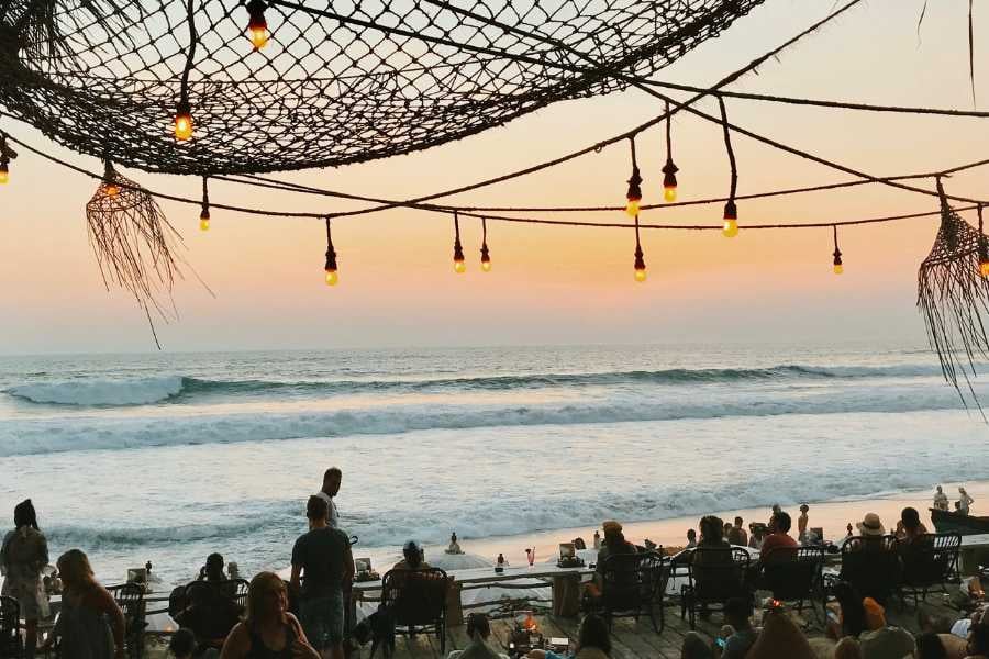 Sunset vibes with string lights, waves, and a relaxed beach crowd.