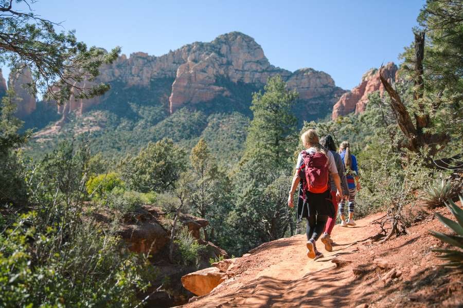 Three hikers follow a dirt trail through the forest with tall red mountains ahead.