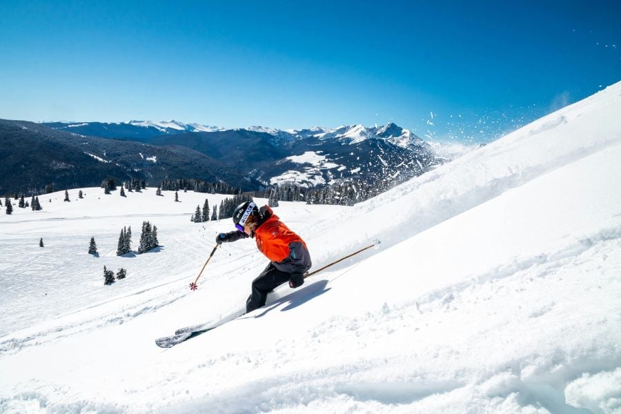Skier racing downhill, carving fresh powder with mountain peaks in the background.