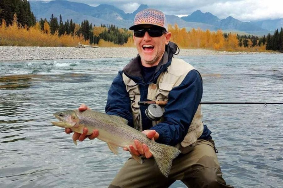 A happy fisherman holding a big trout with mountains and trees behind him.
