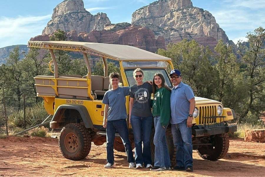 Smiling family poses by a bright yellow jeep with red rock cliffs behind them.