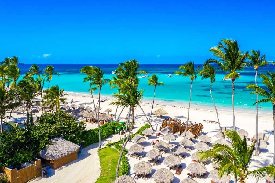 A stunning view of a tropical beach with clear turquoise water, palm trees, and thatched umbrellas.