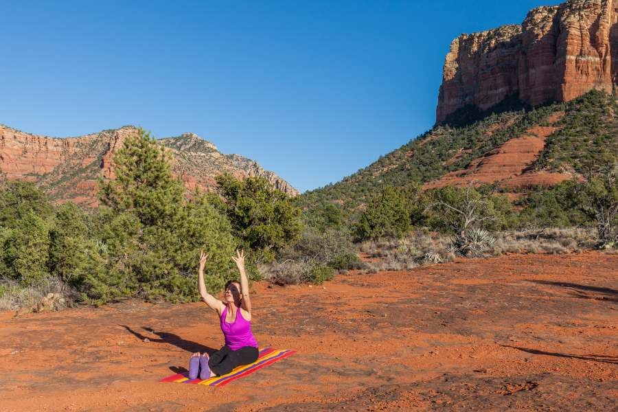 A woman is doing yoga on red earth, finding spiritual peace with mountains and trees behind her.
