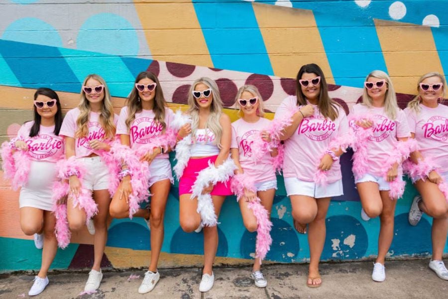 A group of friends dressed in pink Barbie outfits smiles in front of a colorful wall.