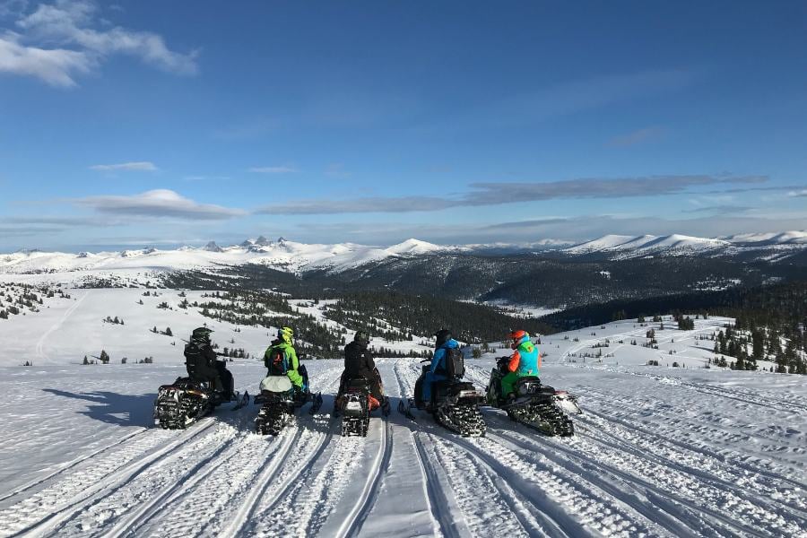 nowmobilers pause on a mountain ridge, overlooking endless snowy peaks.