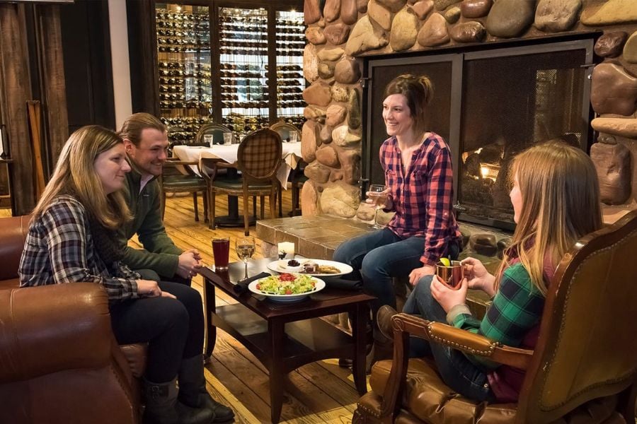 Friends share drinks and laughter by a cozy stone fireplace.