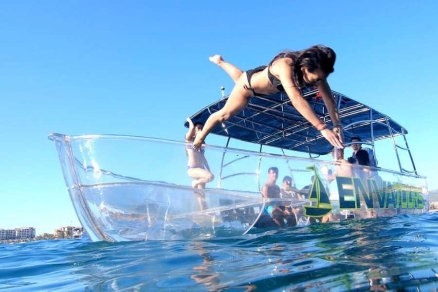 A woman dives off a see-through boat into crystal-clear ocean waters, surrounded by happy friends.