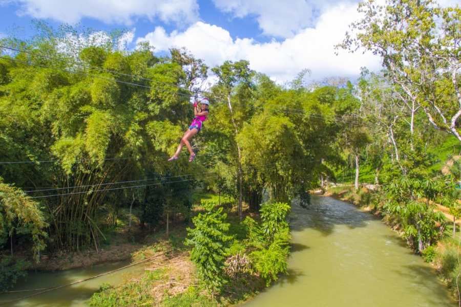 A thrill-seeker glides across a zipline above a winding river, surrounded by towering green bamboo and jungle trees.