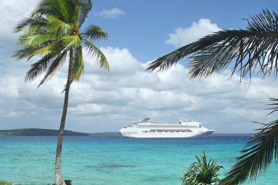 A cruise ship docked in a tropical location, with palm trees lining the beach and tourists walking along the shore.