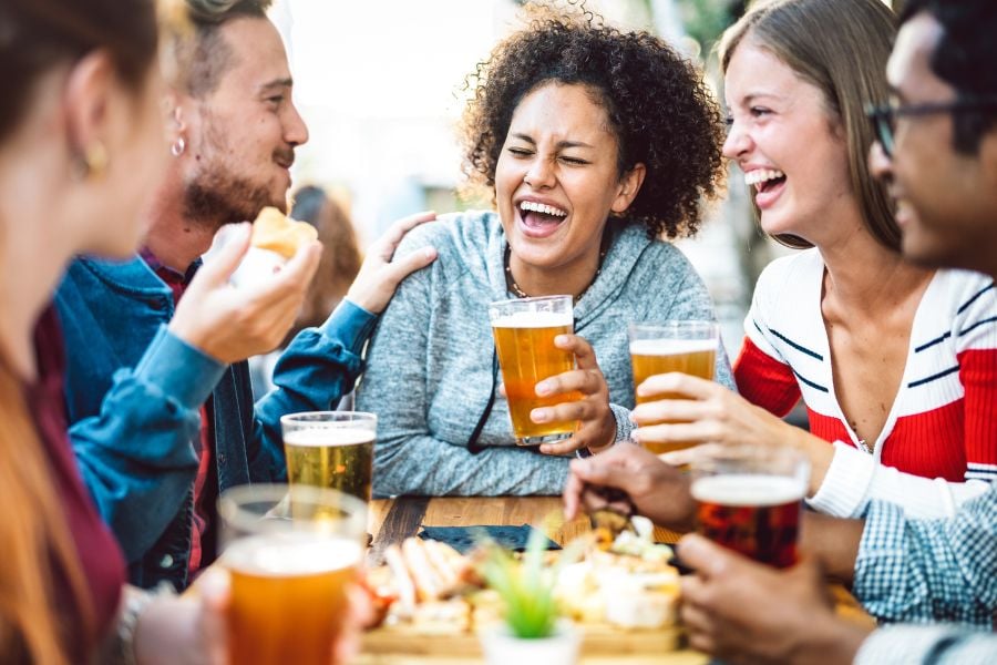 Friends laugh together while enjoying cold beers at an outdoor table.