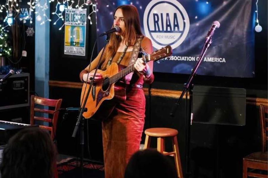 A young woman sings and plays guitar under twinkling lights on stage.