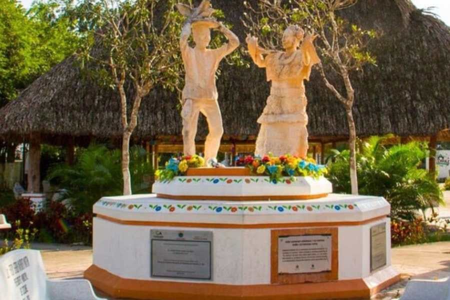 A vibrant statue of a traditional dancing couple, celebrating local culture, surrounded by flowers and thatched-roof buildings.