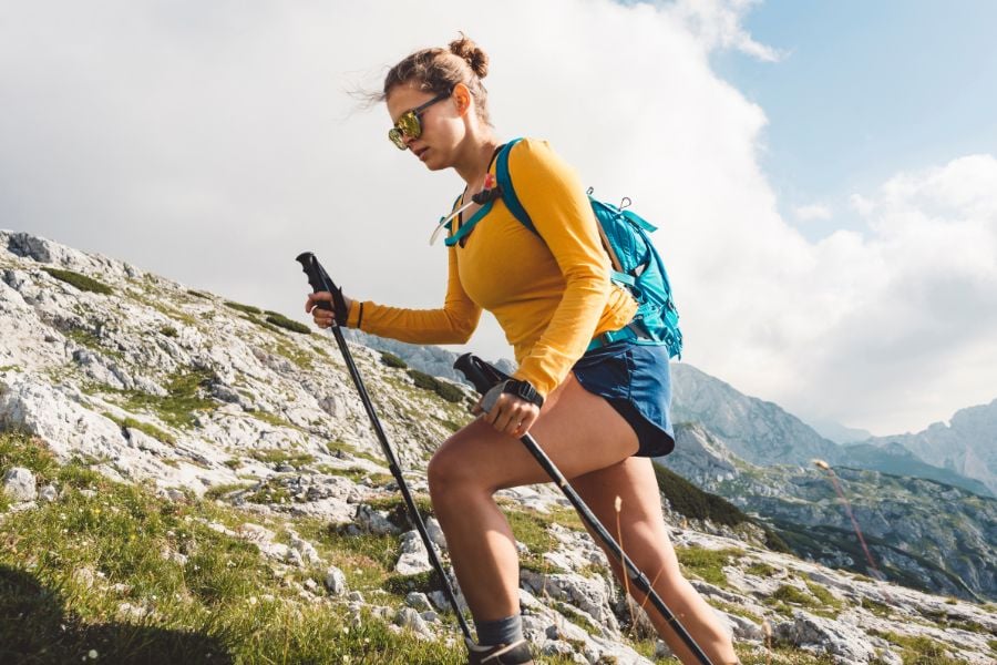 A woman hikes uphill with trekking poles under bright mountain skies.