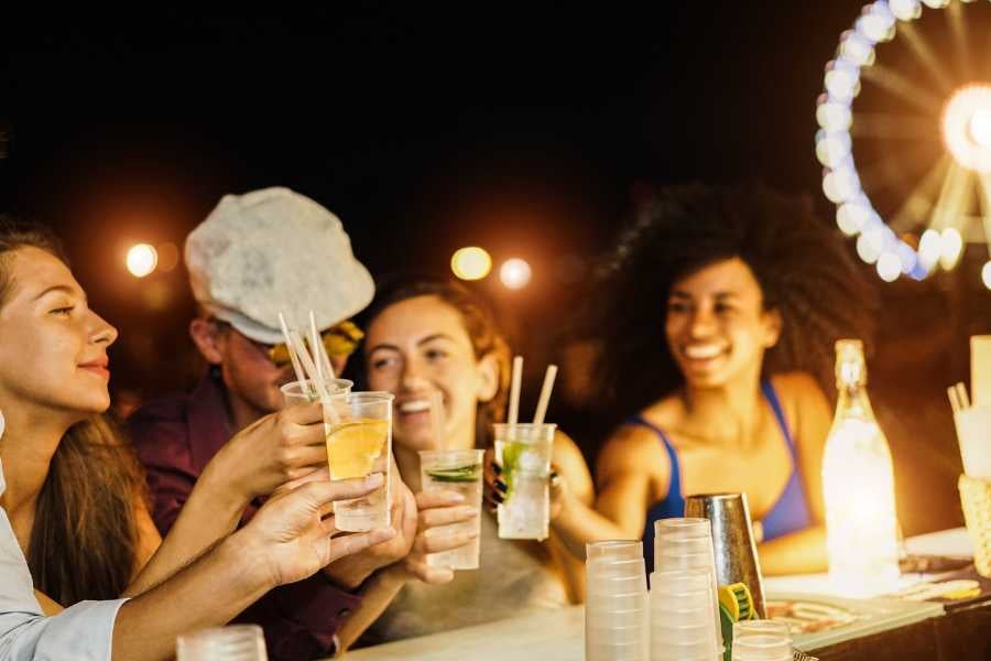 Friends toast with cocktails at a beach bar, with a Ferris wheel glowing in the background.