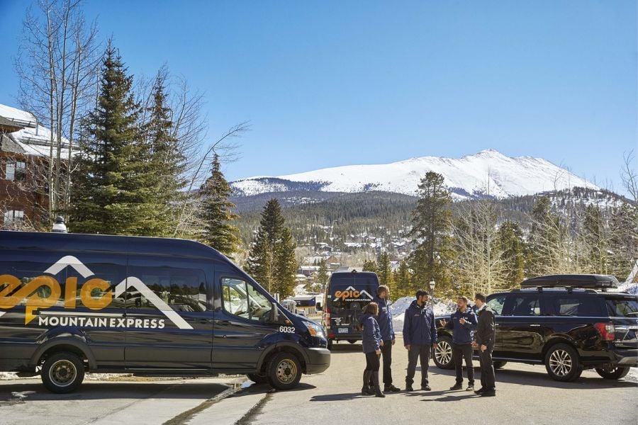 Shuttle vans wait as travelers chat with snowy mountains in the background.