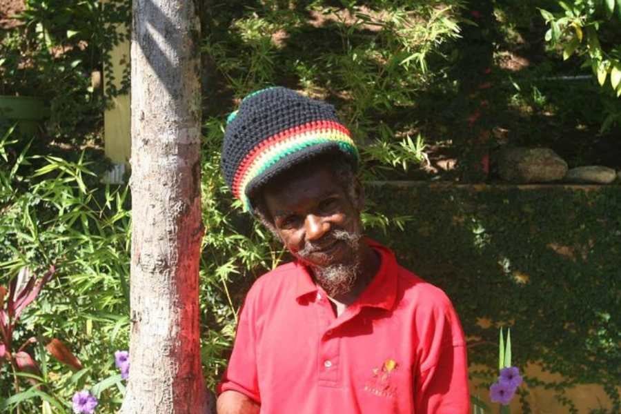 A friendly local man in a colorful hat smiles warmly, leaning against a tree in a lush, tropical setting.