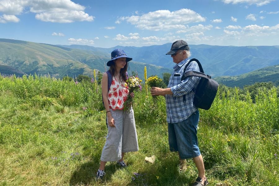 Two people share wildflowers while admiring the rolling hills and blue mountain scenery.