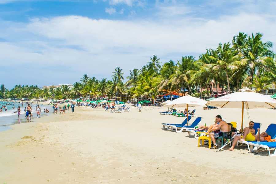 A lively beach filled with people sunbathing and playing in the sand, with palm trees in the background.