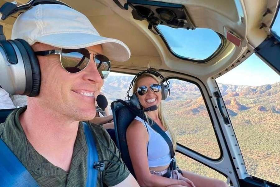 Two people smile while enjoying a scenic helicopter ride over rugged mountain views.