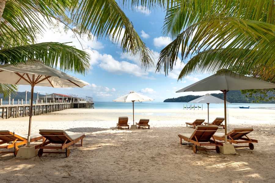 A peaceful beach scene with lounge chairs under umbrellas and a wooden pier in the distance.