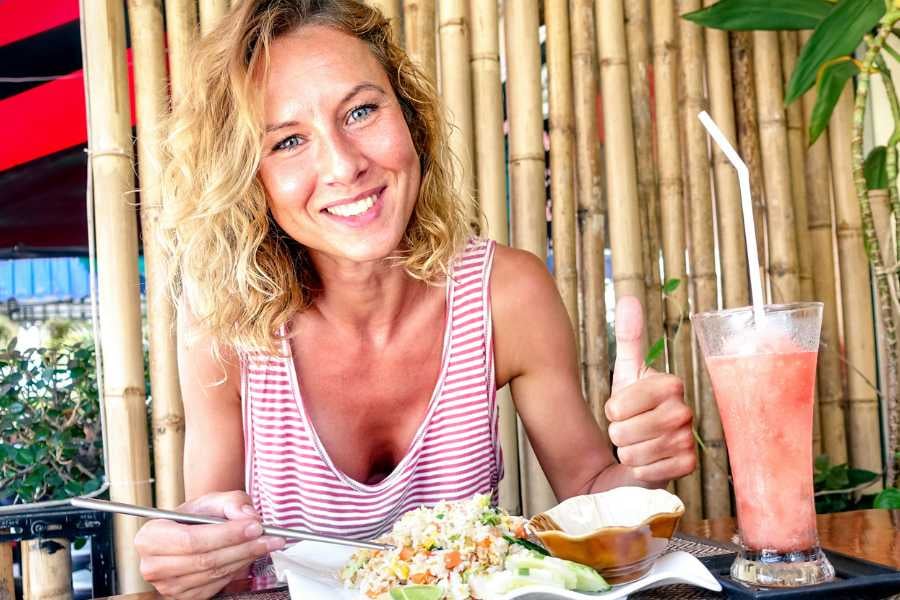 A joyful woman gives a thumbs-up while enjoying a vibrant salad and refreshing pink drink at a bamboo-themed cafe.