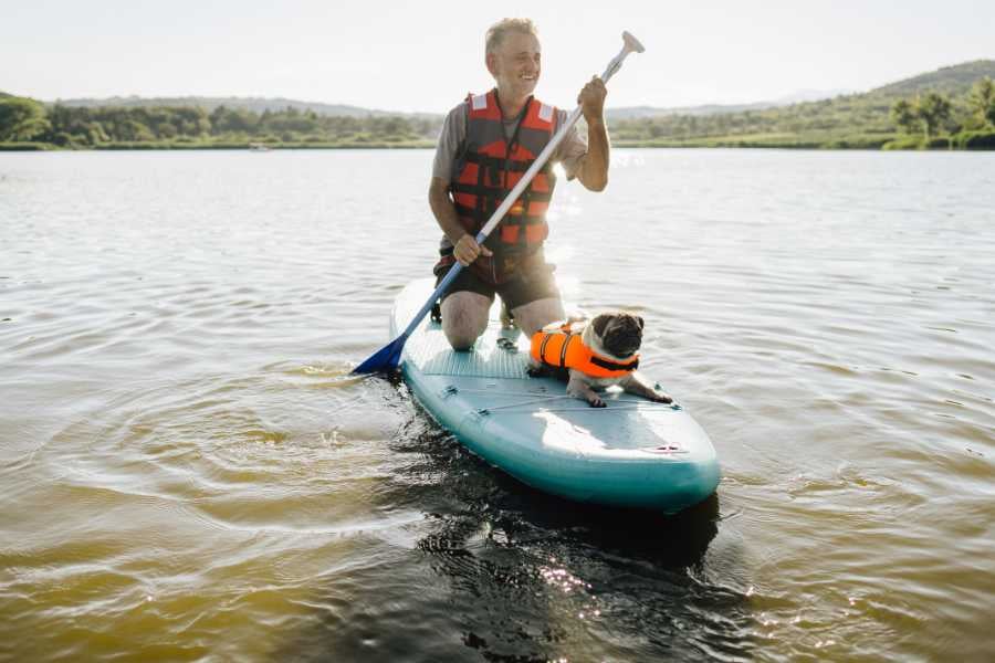 A man paddles on a calm lake with his small dog, both wearing safety vests under the sunny sky.