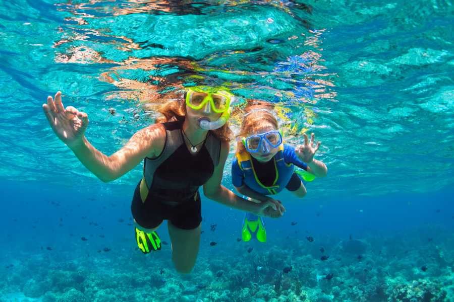  woman and child snorkel together, signaling "OK" above a coral reef.