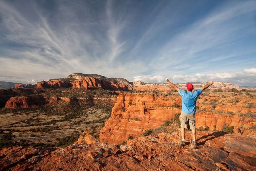  A man celebrates on top of a cliff, overlooking wide red rock landscapes.