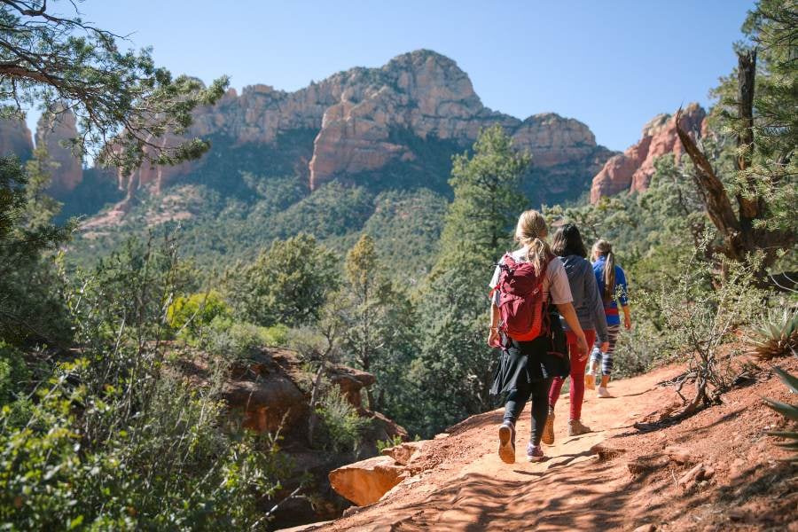 A group hikes a red dirt trail surrounded by pine trees and giant rocky cliffs.