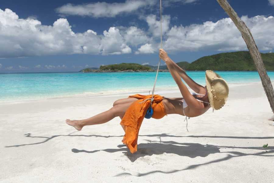 A woman swings under a shady tree on a pristine beach with crystal-clear waters in the background.