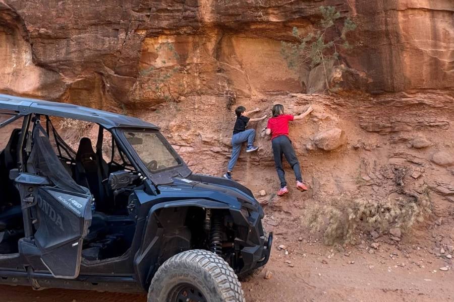 Two kids are climbing a rock wall next to an off-road vehicle.