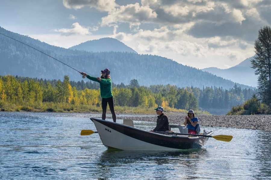 A fly fisher casts into a calm river, with mountains and golden trees in the background.