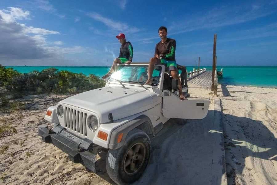 Two adventurers sit on a white Jeep, enjoying ocean views with bright blue waters and a wooden pier nearby.