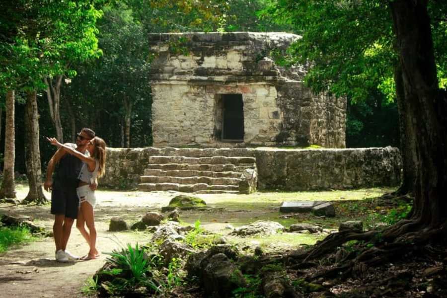 A couple poses happily in front of an ancient Mayan ruin, surrounded by tall trees and dappled sunlight.