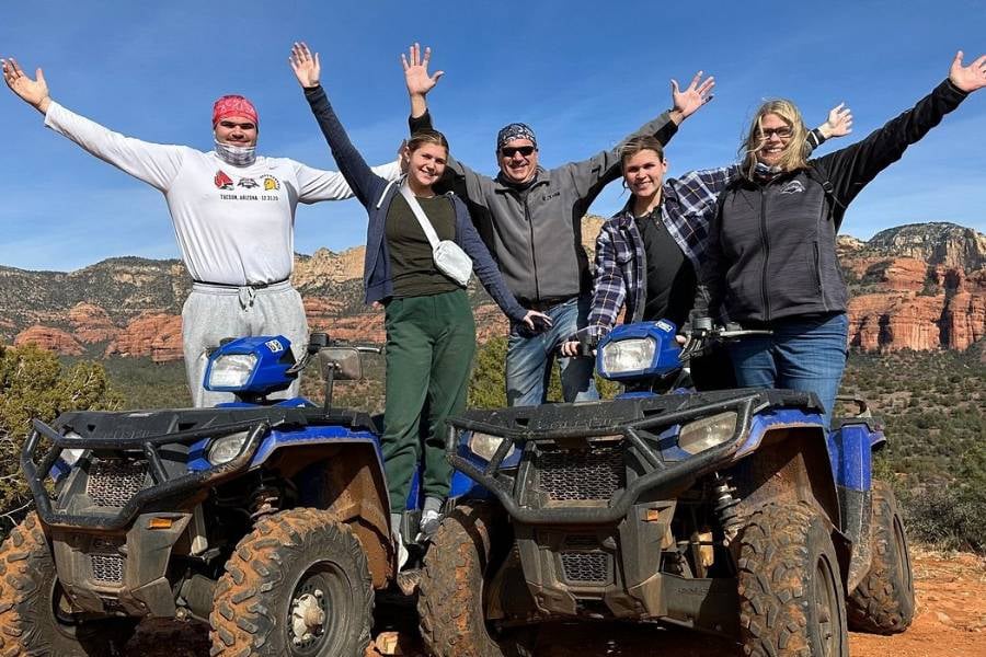 Five people are standing with ATVs, smiling and raising their hands.