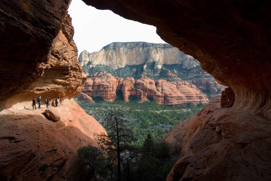 Hikers walk along a cliffside cave with stunning red rock views.