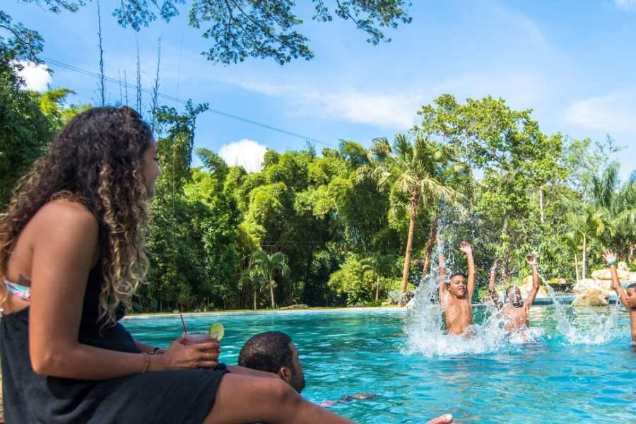 Friends splash and relax in a crystal-clear pool, surrounded by tall palm trees and vibrant green nature.