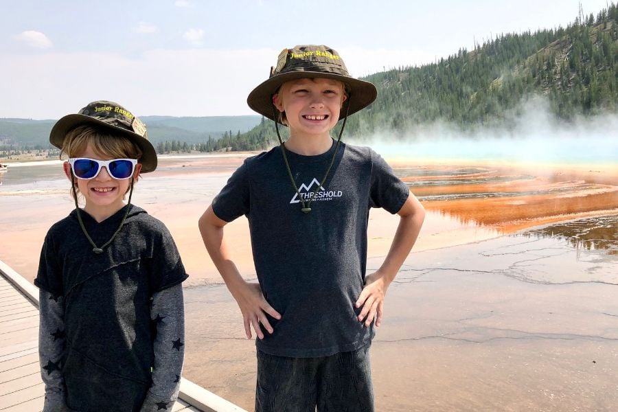 Two kids in ranger hats smile near Yellowstone’s colorful hot spring.