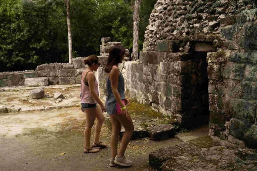 Two women explore ancient ruins, stepping through a moss-covered stone doorway in a peaceful, hidden jungle site.