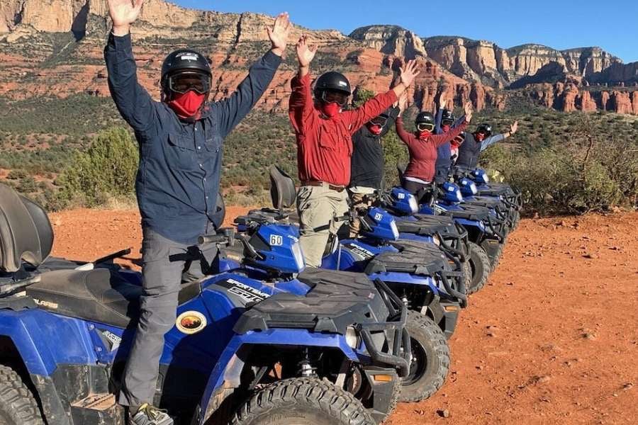 A line of riders stand on their ATVs, raising their arms in celebration.