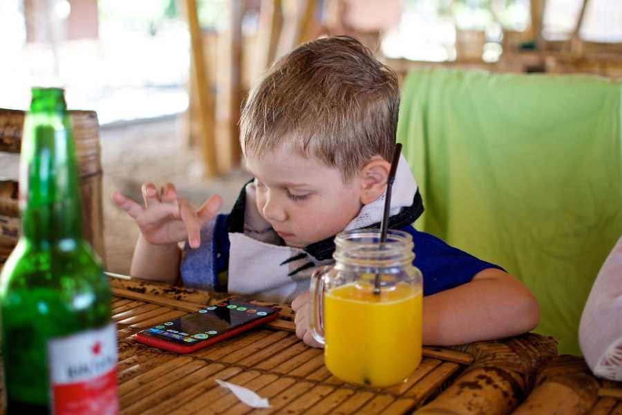 A young boy focused on a phone while sipping fresh orange juice at a cozy, rustic table.