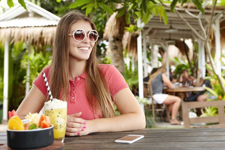 A cheerful woman wearing sunglasses enjoys a tropical drink at a relaxing outdoor cafe.