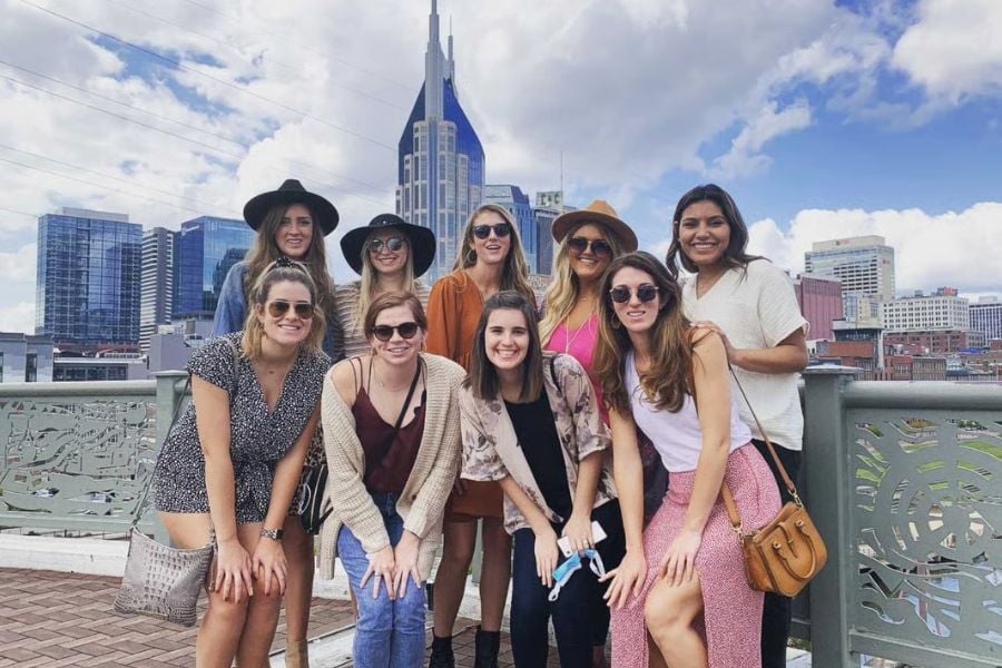 A group of friends smiles together in front of city buildings and a blue sky.